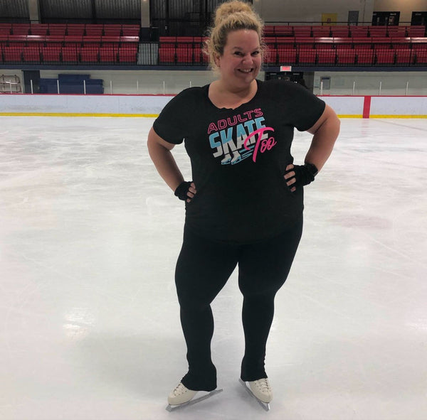 A smiling skater in a black Adults Skate Too tee with her hands on her hips on an indoor rink.