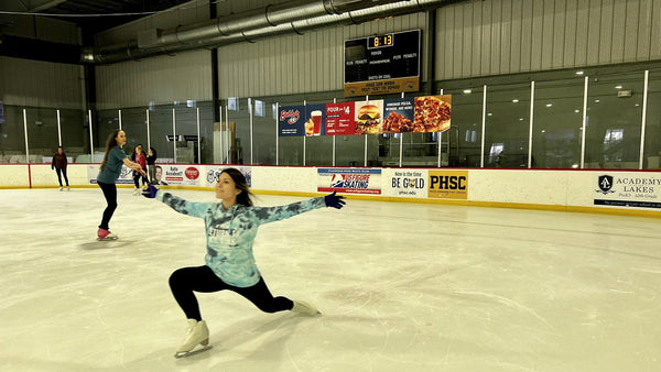 An adult skater gliding in a deep lunge with her arms out while another skater practices behind her