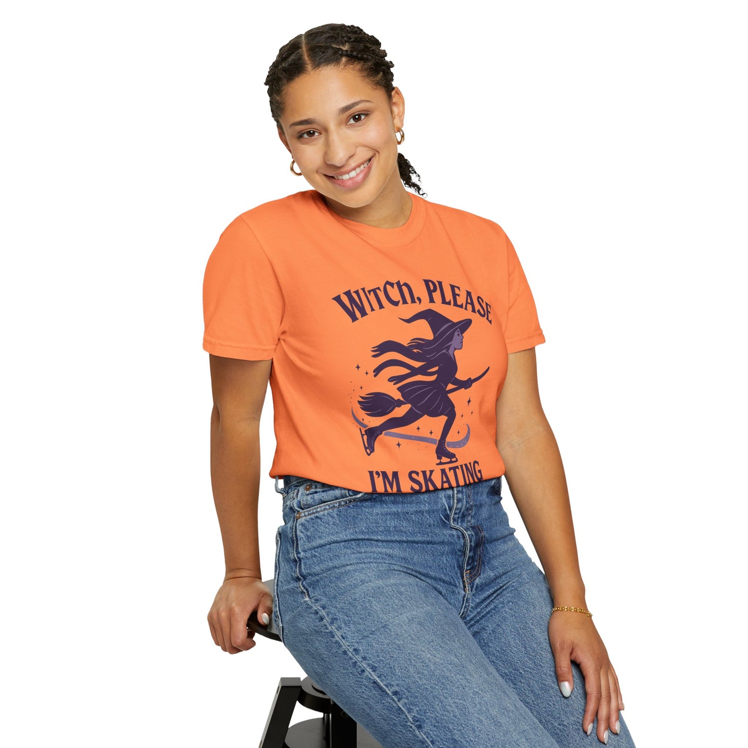 Person wearing an orange t-shirt with a witch graphic and text, sitting on a stool against a white background.