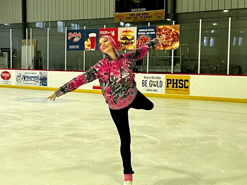 An adult skater in a pink camo hoodie balancing on one foot on the ice with her arms out and her free leg bent behind her