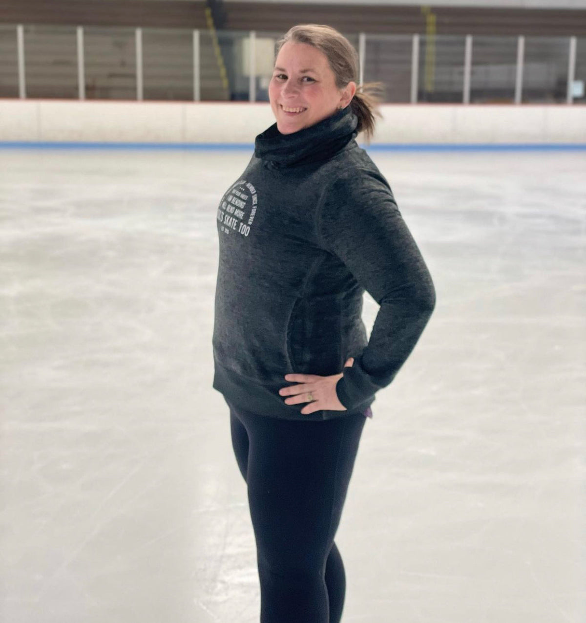 A skater in a grey cowl-neck Adults Skate Too sweatshirt standing on an indoor rink.