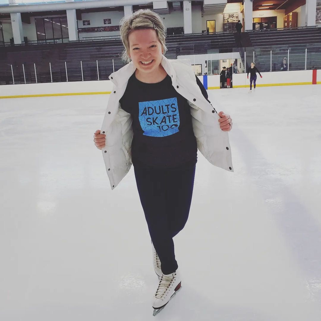 A smiling skater opening a white jacket to show an Adults Skate Too tee on an indoor rink.