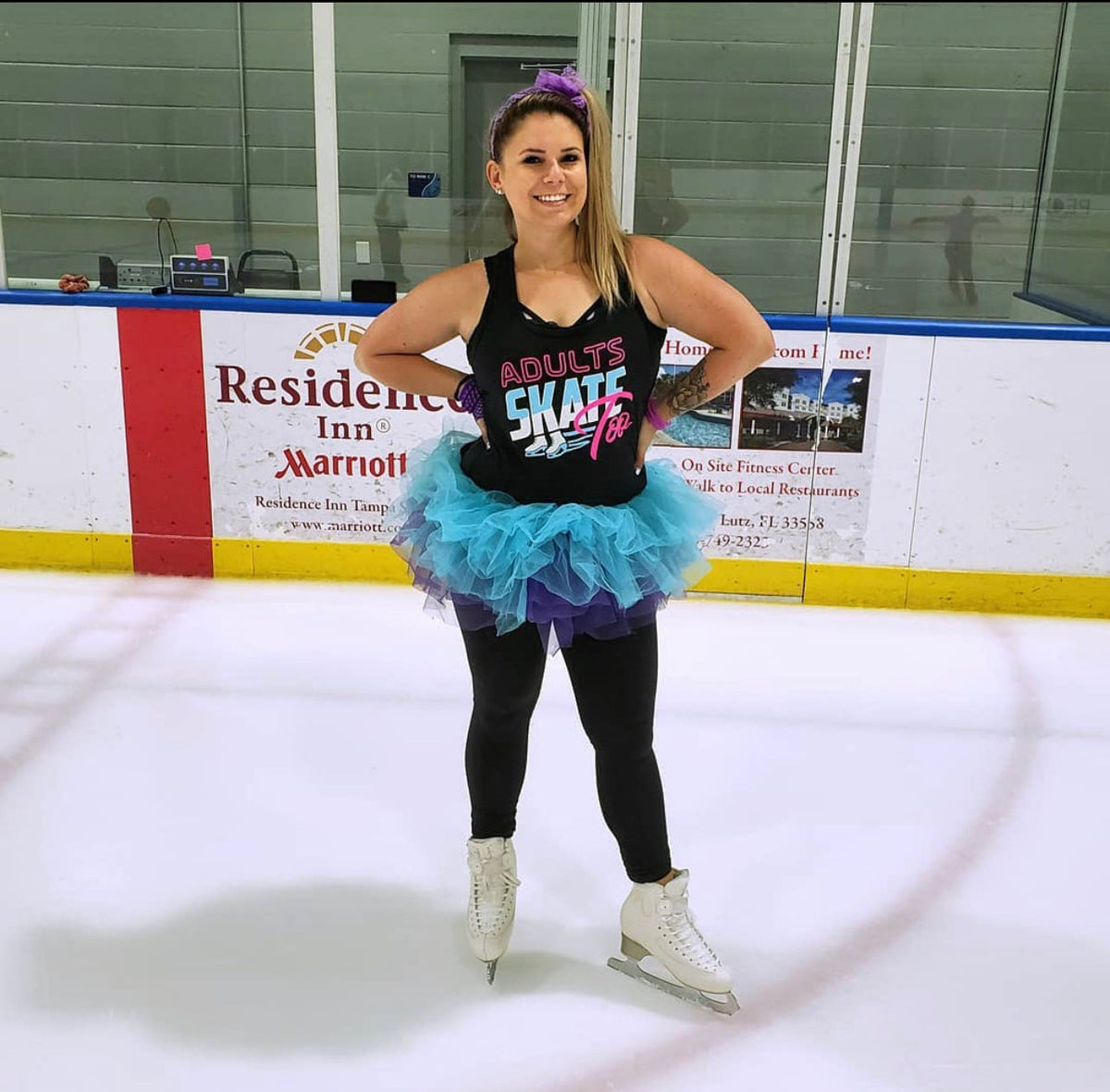 An adult skater in an Adults Skate Too tank top and a blue and purple tutu smiling on the ice in white figure skates