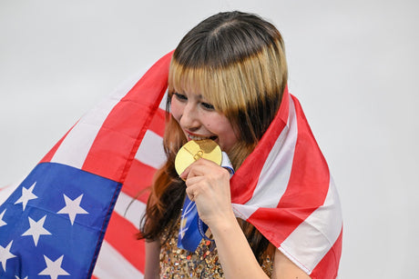 Alysa Liu holds her gold medal to her teeth with the U.S. flag around her shoulders at the 2026 Winter Olympics in Milano Cortina.
