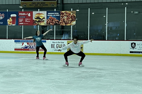 Two adult skaters practising on a quiet rink, one with her arms out and one in a deep knee bend