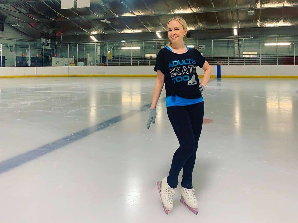 An adult skater in an Adults Skate Too tee standing on the ice at an indoor rink with one hand on her hip