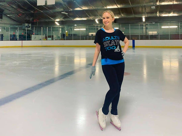 An adult skater in an Adults Skate Too tee standing on the ice at an indoor rink with one hand on her hip
