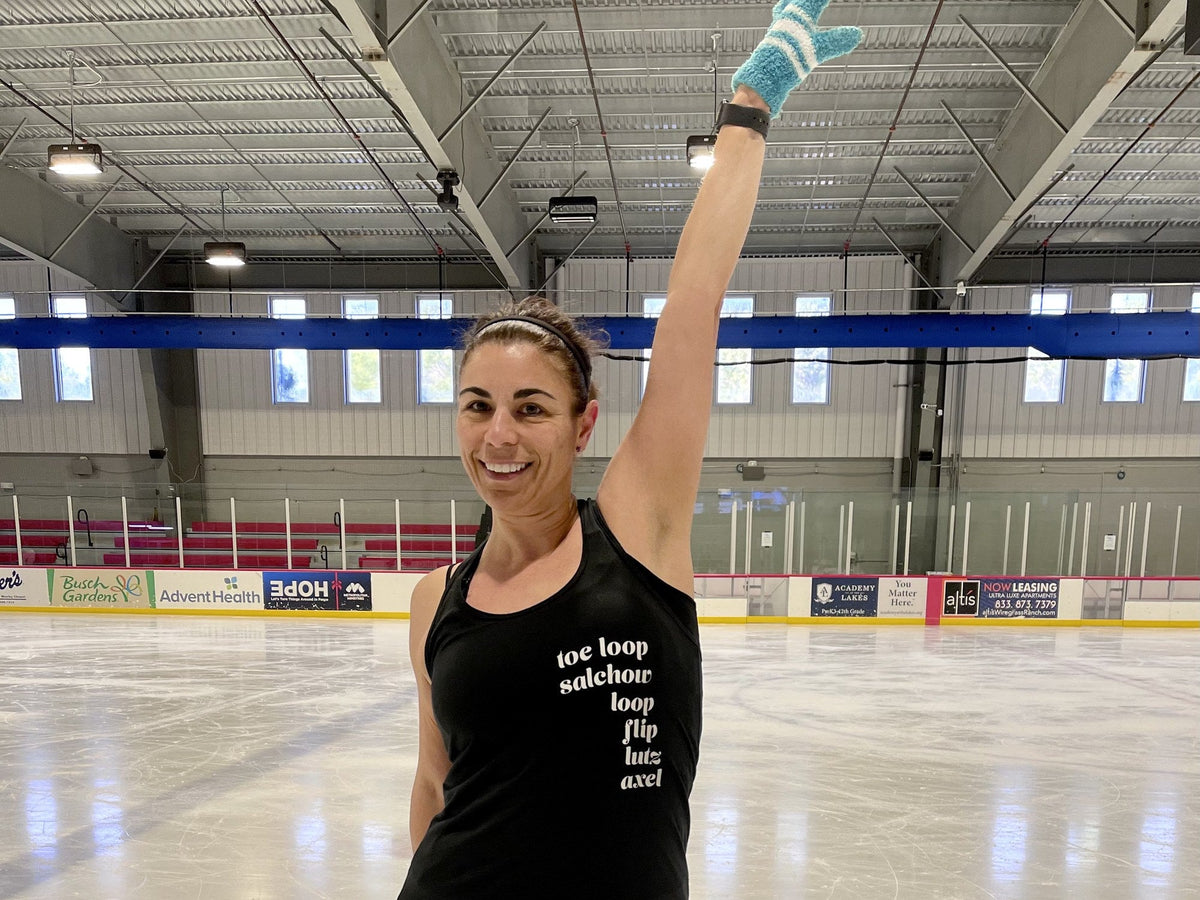 An adult skater smiling on the ice with one skate held up high beside her head