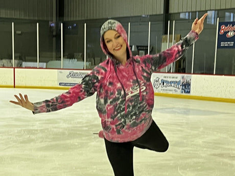 An adult skater in a pink tie-dye hoodie balancing on one foot on the ice, arms out wide and smiling
