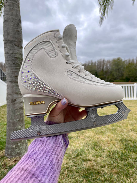 A hand holding up a white figure skate with crystal detailing outdoors, the blade's curve showing.