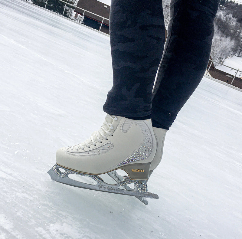 A skater's white figure skate with a crystal-studded blade standing on outdoor ice.