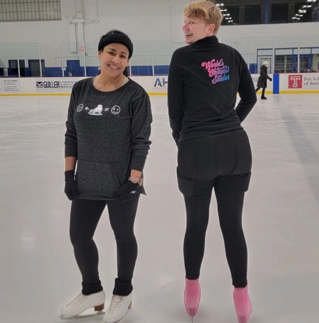 Two skaters chatting on an indoor rink, one showing the back of a World's Okayest Skater jacket.