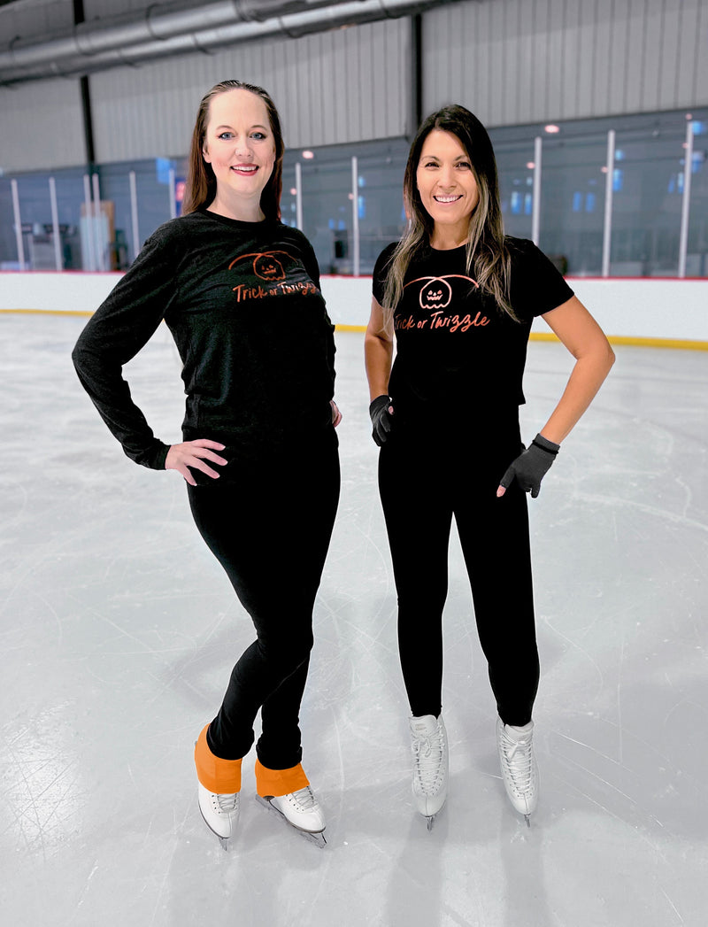 Two skaters in black Trick or Twizzle tees standing on an indoor rink.