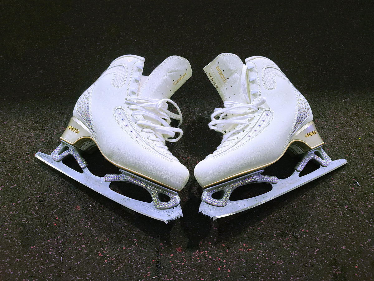 A pair of white figure skates with crystal detailing on a black floor.