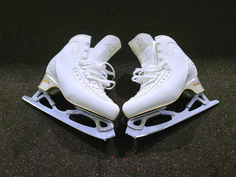 A pair of white figure skates with crystal detailing on a black floor.