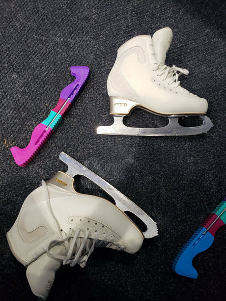 Two white figure skates and pink and blue blade guards on a dark rubber floor.