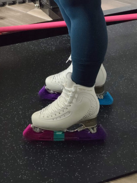 A skater's legs in white figure skates with pink and purple blade guards.