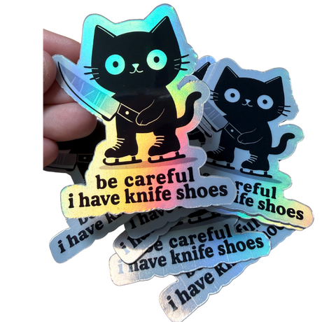 Holographic sticker with a black cat and text 'be careful I have knife shoes' held by a hand.