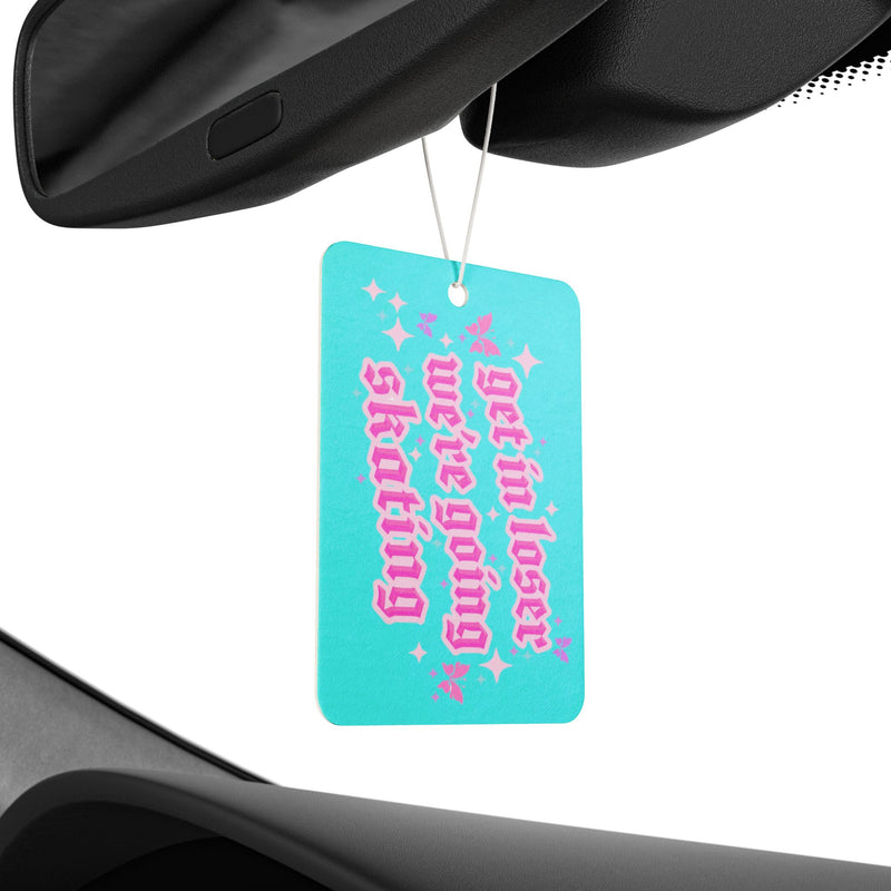 Turquoise car air freshener with pink text hanging from a car's rearview mirror.