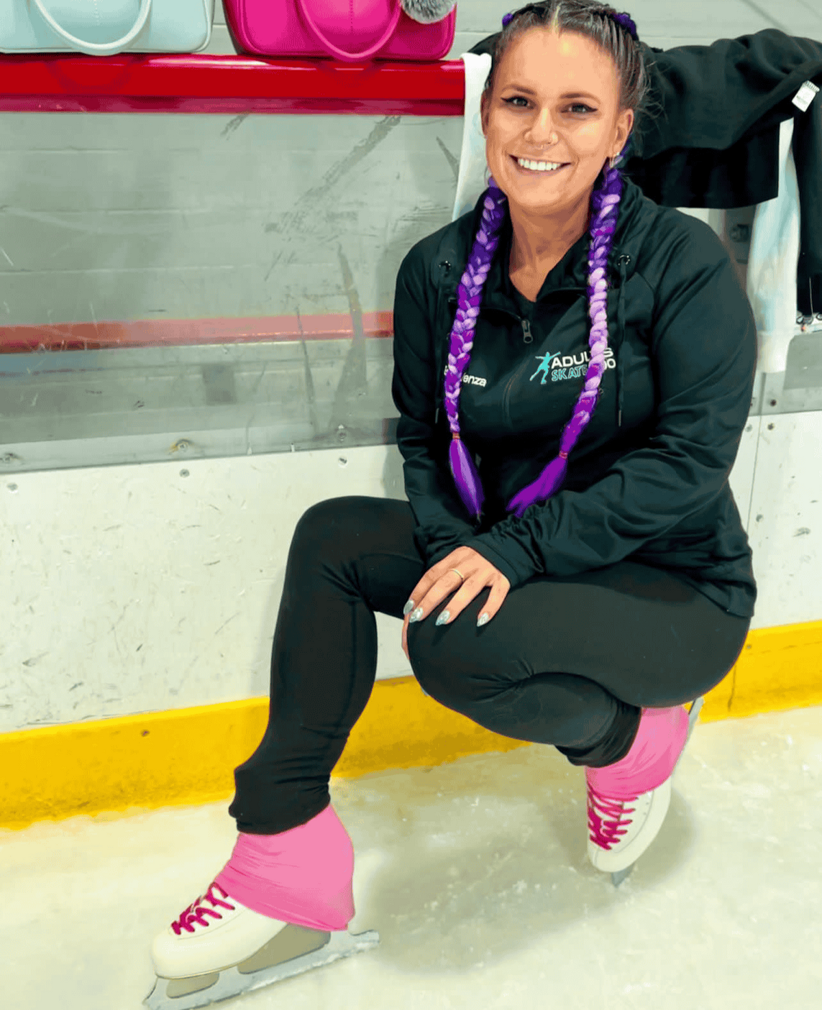 Erika crouching on the ice by the rink boards in her Adults Skate Too jacket and pink boot covers