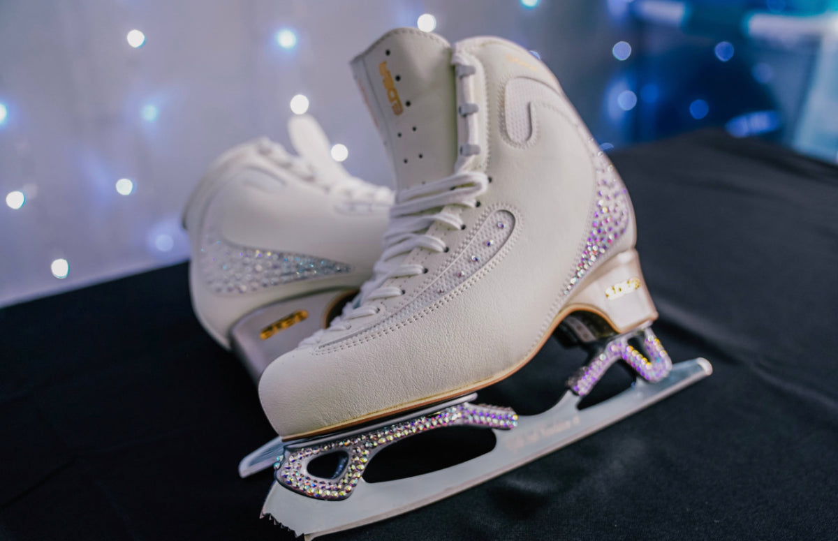 White figure skates with crystal-studded blades on a black cloth with sparkling lights behind.