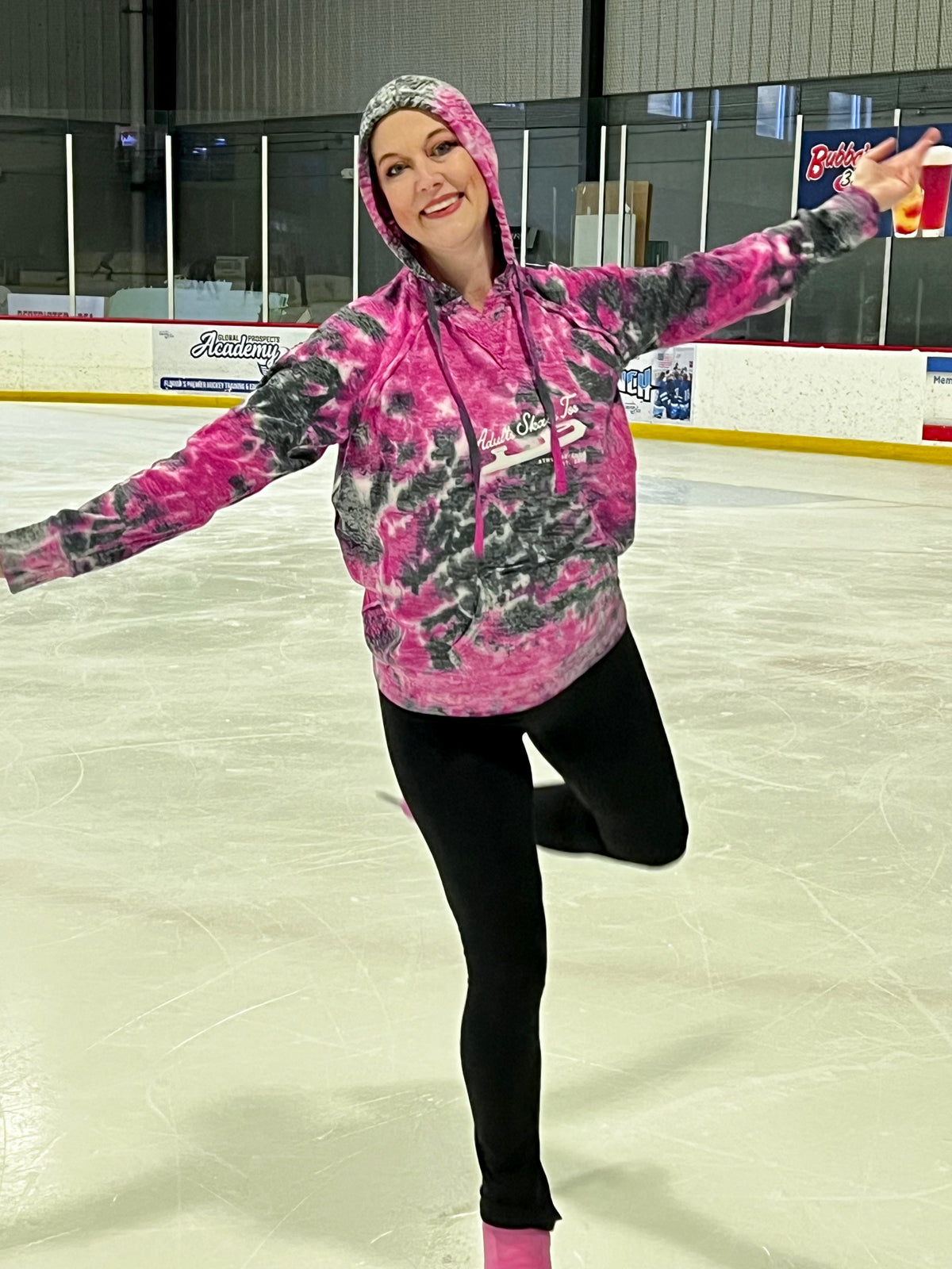 A skater in a pink camo hoodie balancing on one foot with her arms out on an indoor rink.