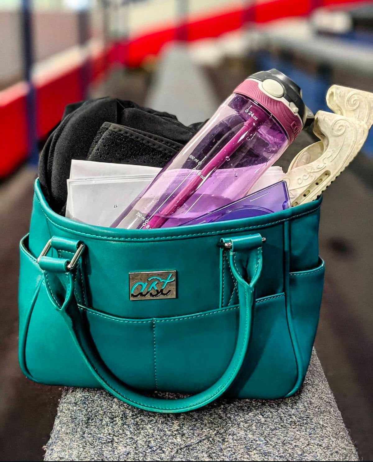 A teal Adults Skate Too tote packed with a water bottle, gloves and blade guards beside the rink.