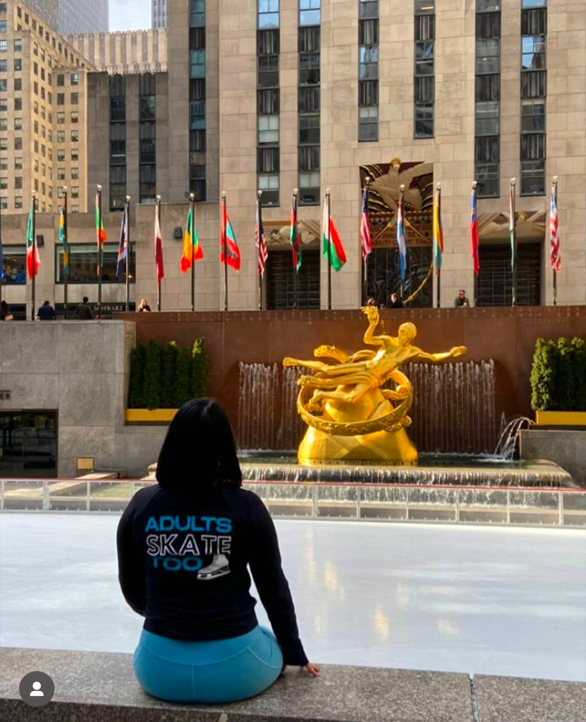 A skater in an Adults Skate Too hoodie sitting by the Rockefeller Center rink with flags behind the gold statue.
