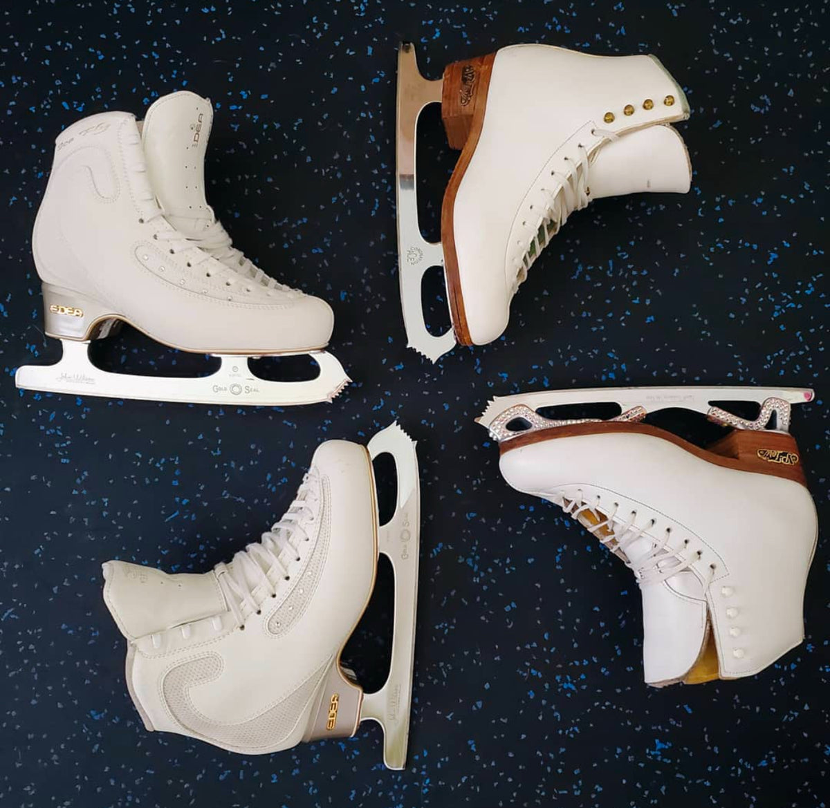 Two pairs of white figure skates laid out on a speckled rubber floor.
