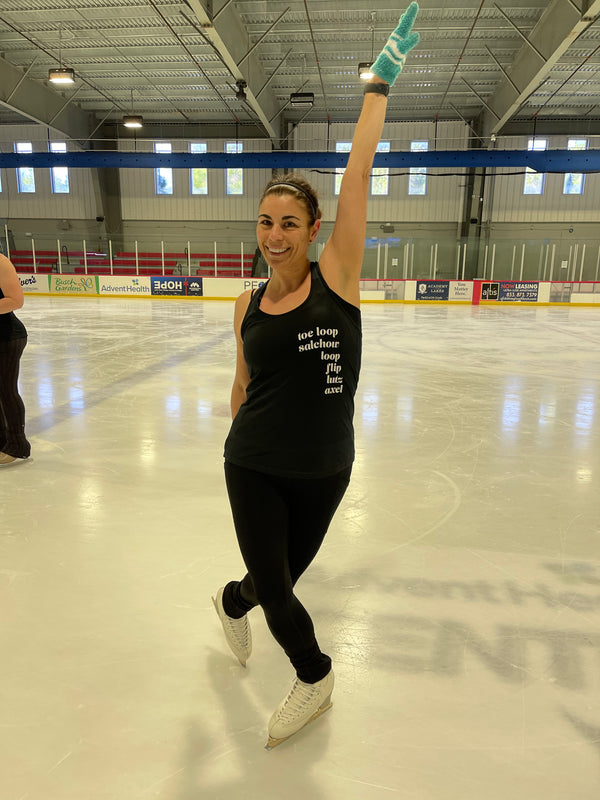 A smiling skater in a black tank printed with jump names, one arm raised, on an indoor rink