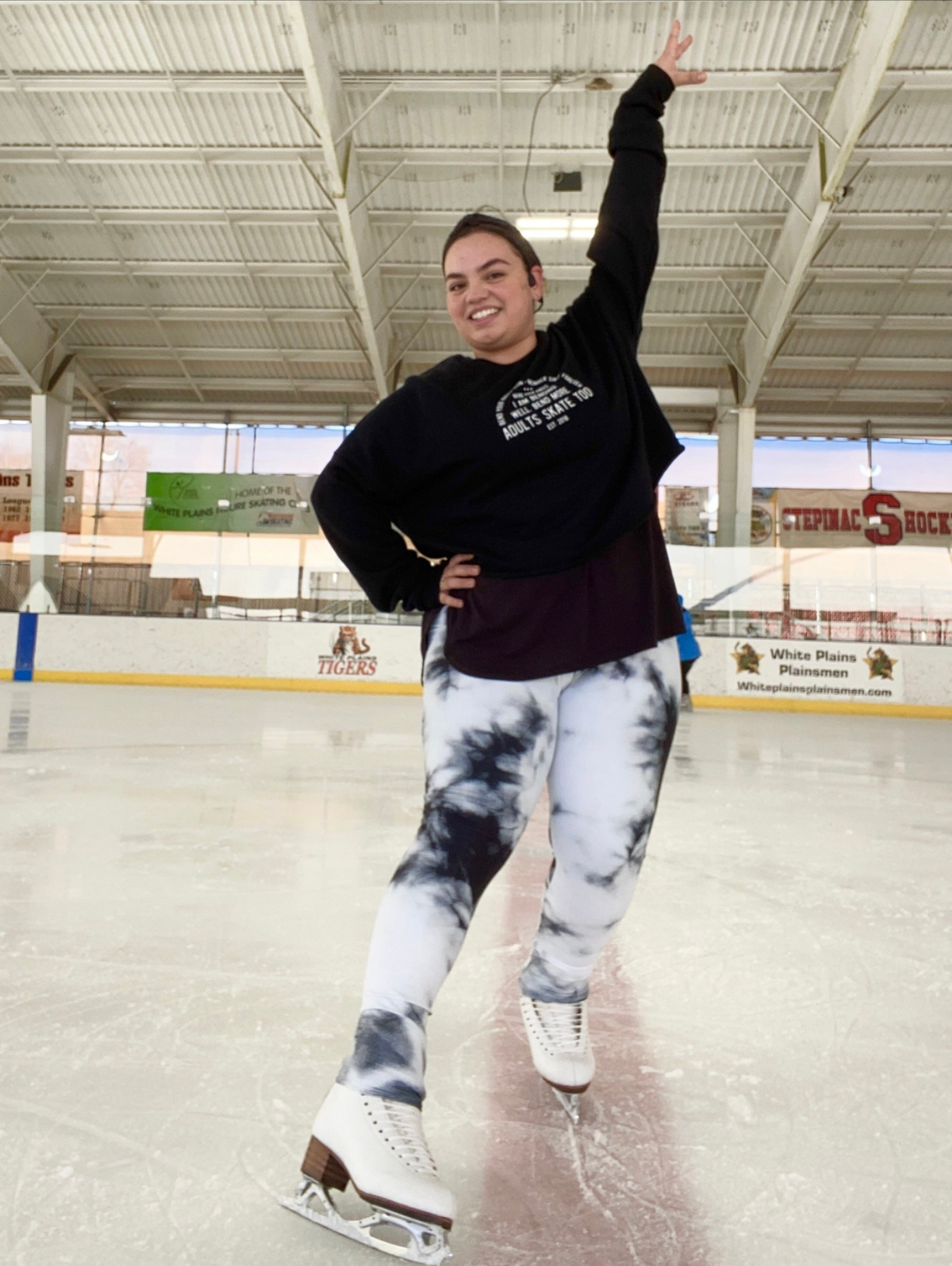 A skater on practice ice with one arm raised, in a black Adults Skate Too sweatshirt and tie-dye leggings.