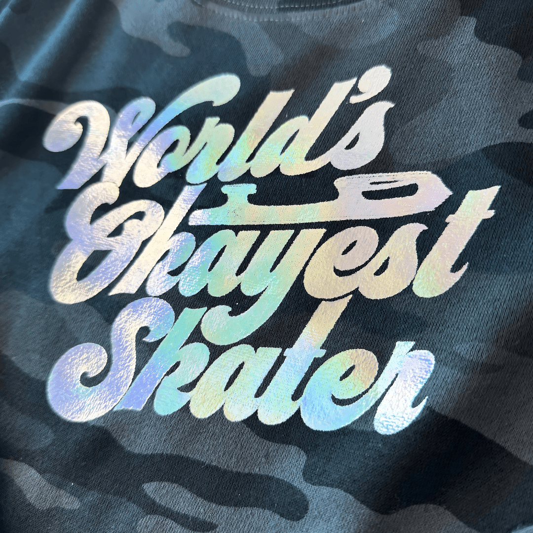 Holographic 'World's Okayest Skater' text on a camouflage-patterned shirt.