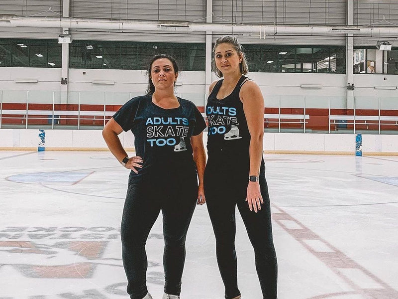 Two adult skaters in Adults Skate Too tees standing on the ice at a rink