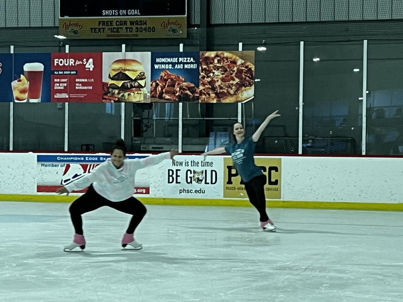 Two adult skaters on a practice session, the rink scoreboard behind them