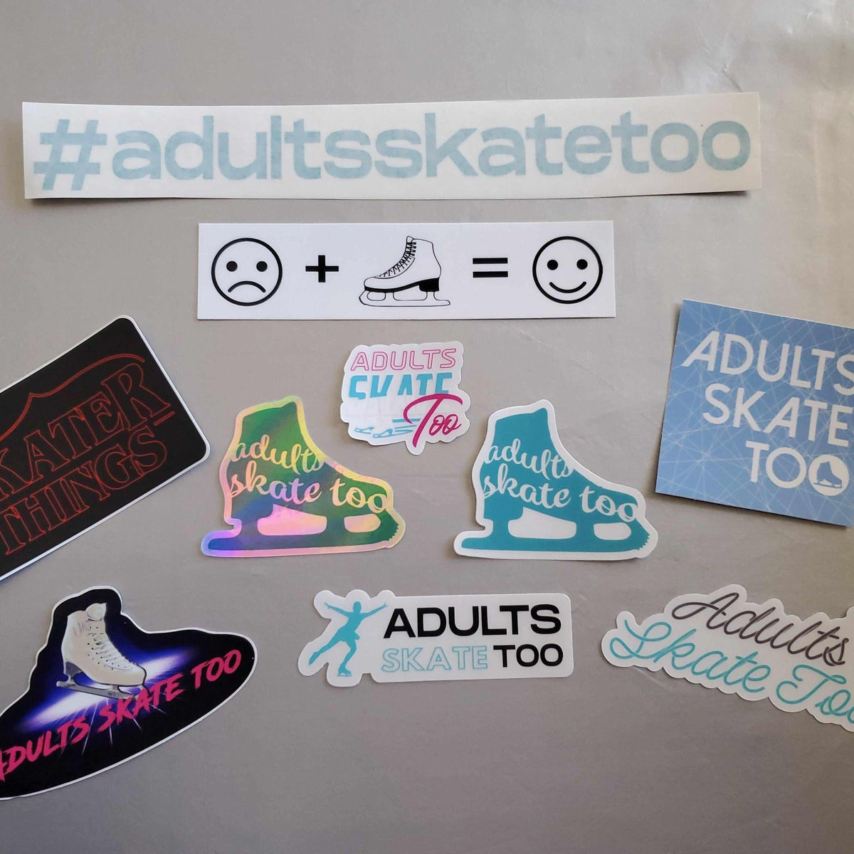 Assorted Adults Skate Too vinyl stickers laid out on a grey table, with the #adultsskatetoo banner across the top