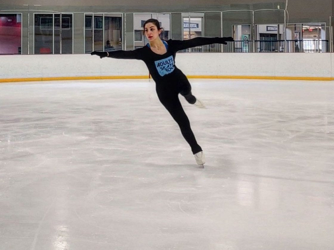 An adult skater in an Adults Skate Too tee gliding on one foot with her arms out on an empty rink