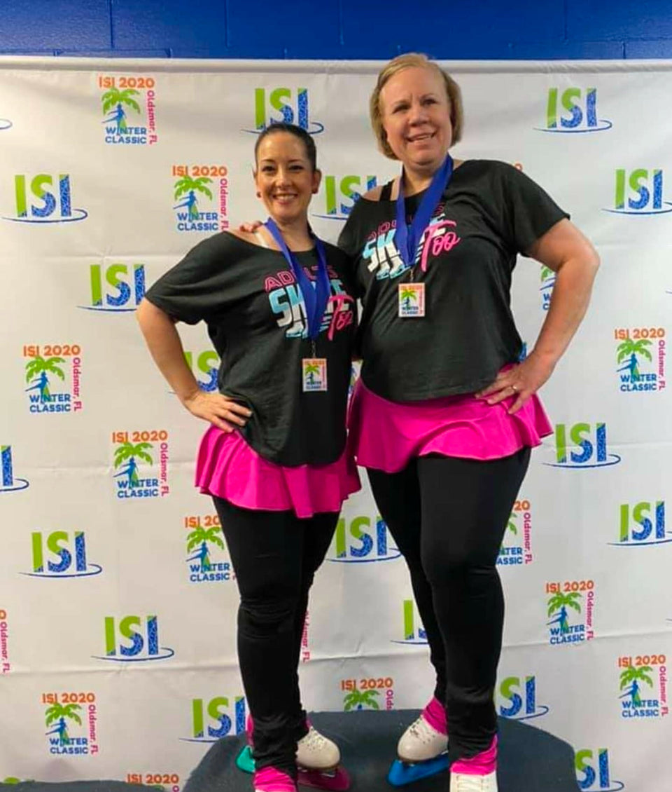 Two adult skaters in Adults Skate Too tees and pink skirts wearing their medals in front of an ISI Winter Classic backdrop