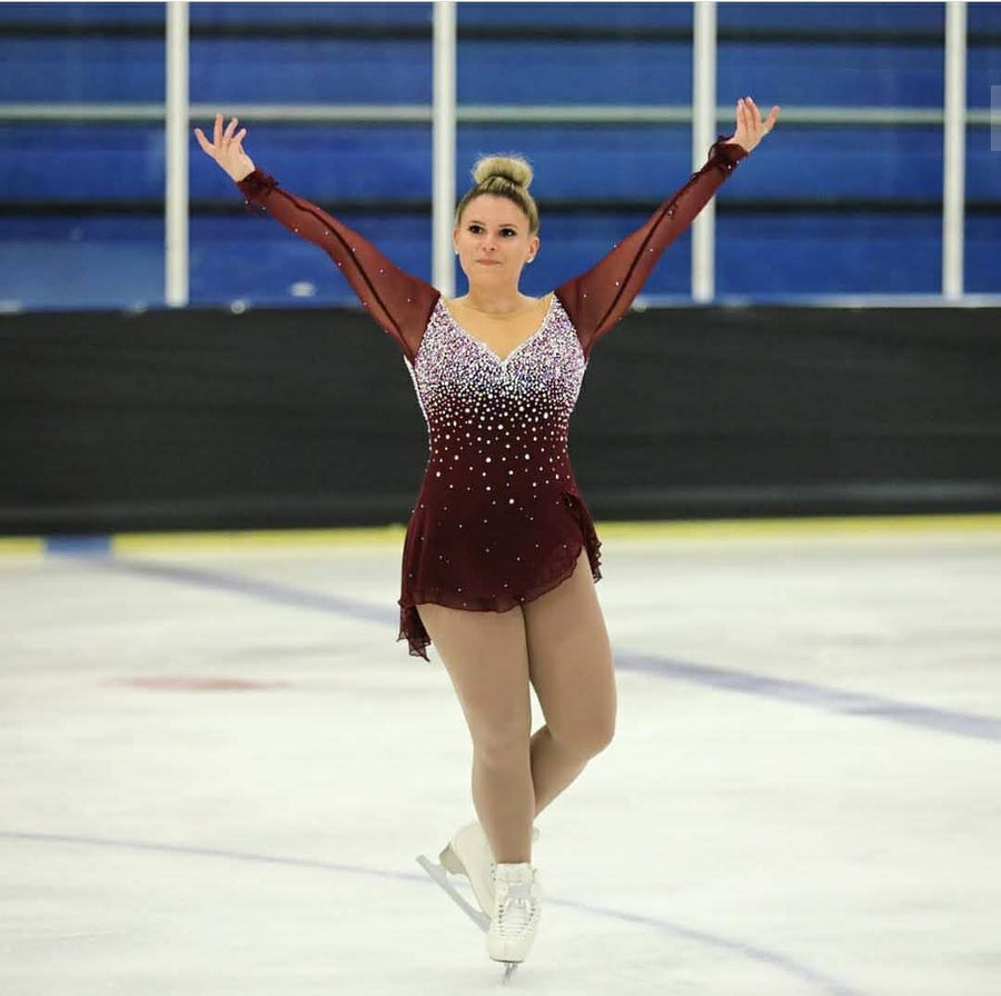 A skater in a burgundy competition dress with both arms raised on the ice