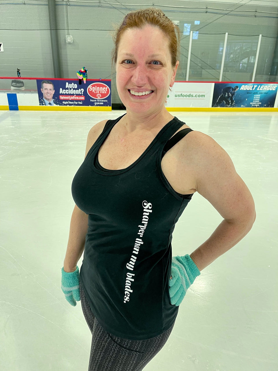 A smiling skater in a black tank with a printed slogan and teal gloves, hands on her hips, on an indoor rink