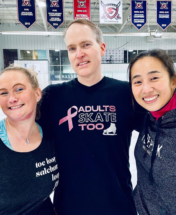 Three smiling skaters at a rink, the man in the middle in a pink ribbon Adults Skate Too tee.