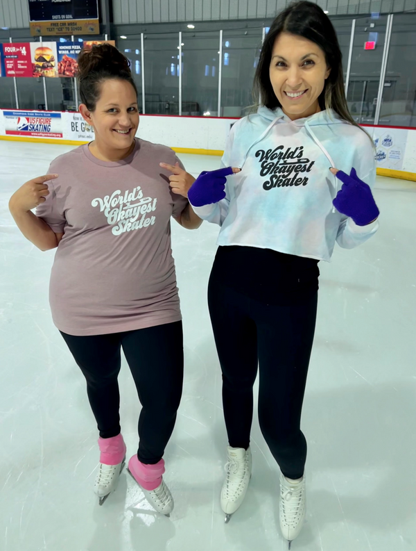 Two smiling skaters in World's Okayest Skater tops on an indoor rink.